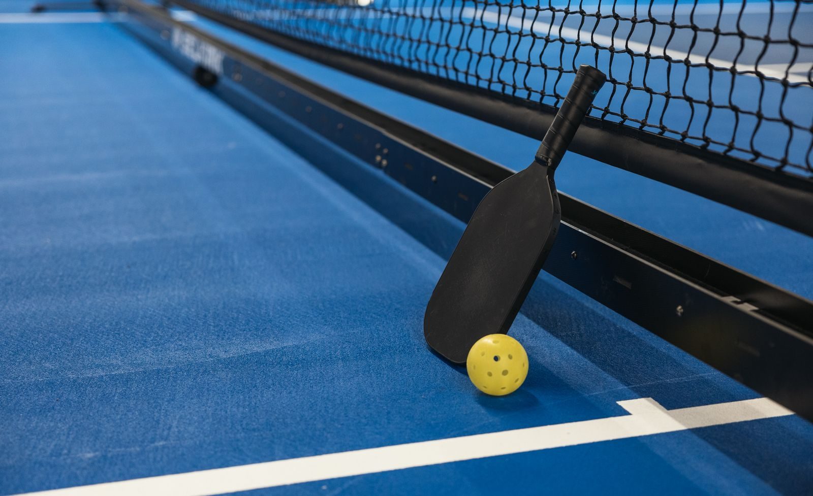 Padel racket and ball resting by the net on a blue padel court