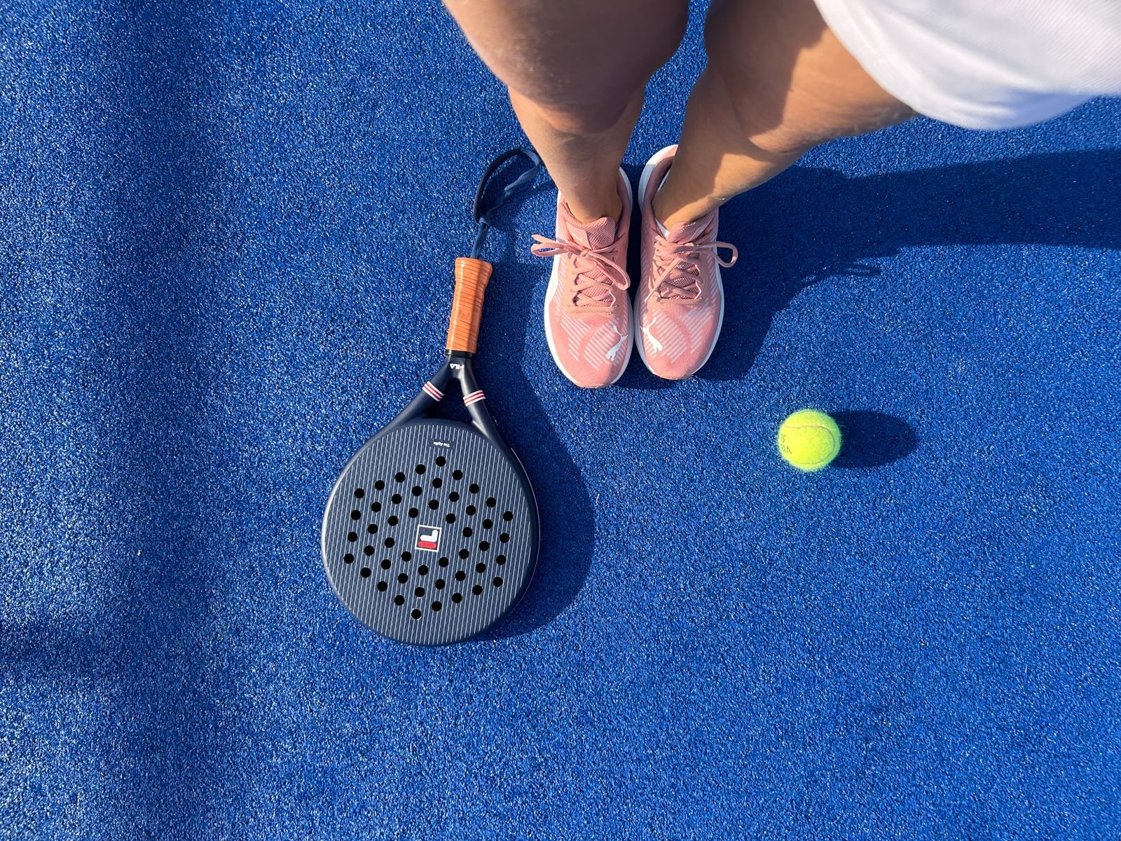 Padel racket and ball on a blue court next to a player's trainers